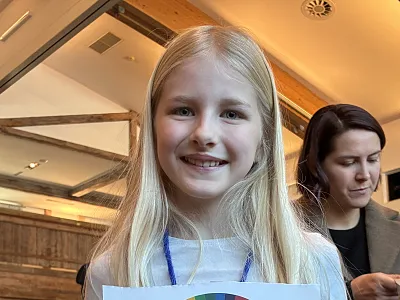 Ein lächelndes Mädchen mit langen blonden Haaren hält ein buntes Plakat in einem hellen Raum. Eine erwachsene Frau steht unscharf im Hintergrund.