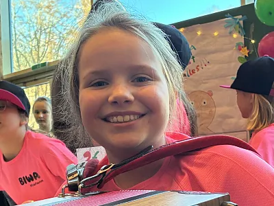 Ein Kind in einem pinken Shirt hält eine Ziehharmonika und lächelt fröhlich in die Kamera. Im Hintergrund sind weitere Kinder in ähnlichen T-Shirts zu sehen.