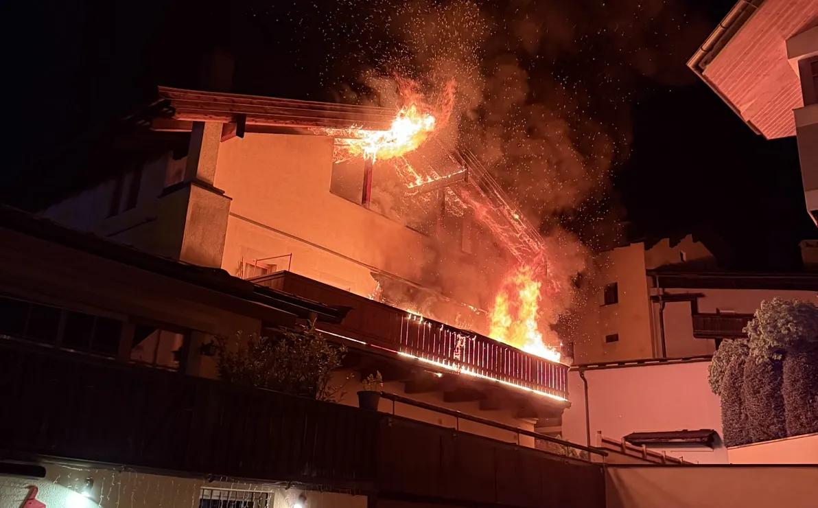 Nachthimmel mit brennendem Dach eines Gebäudes, Flammen und Rauch steigen auf, während umliegende Häuser im Dunkeln bleiben. Dramatik der Szene ist deutlich.