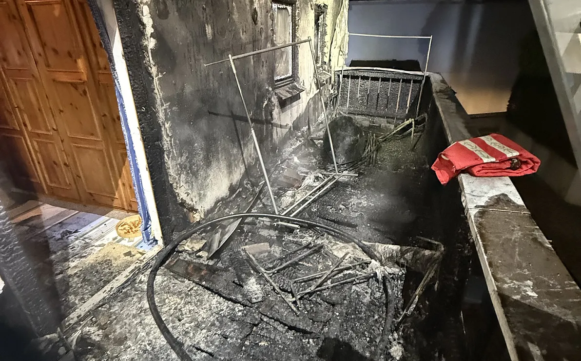 Verbrannter Balkon mit verkohlten Überresten von Möbeln, einem geschmolzenen Geländer und Ruß an den Wänden, deutet auf kürzlichen Brand hin.