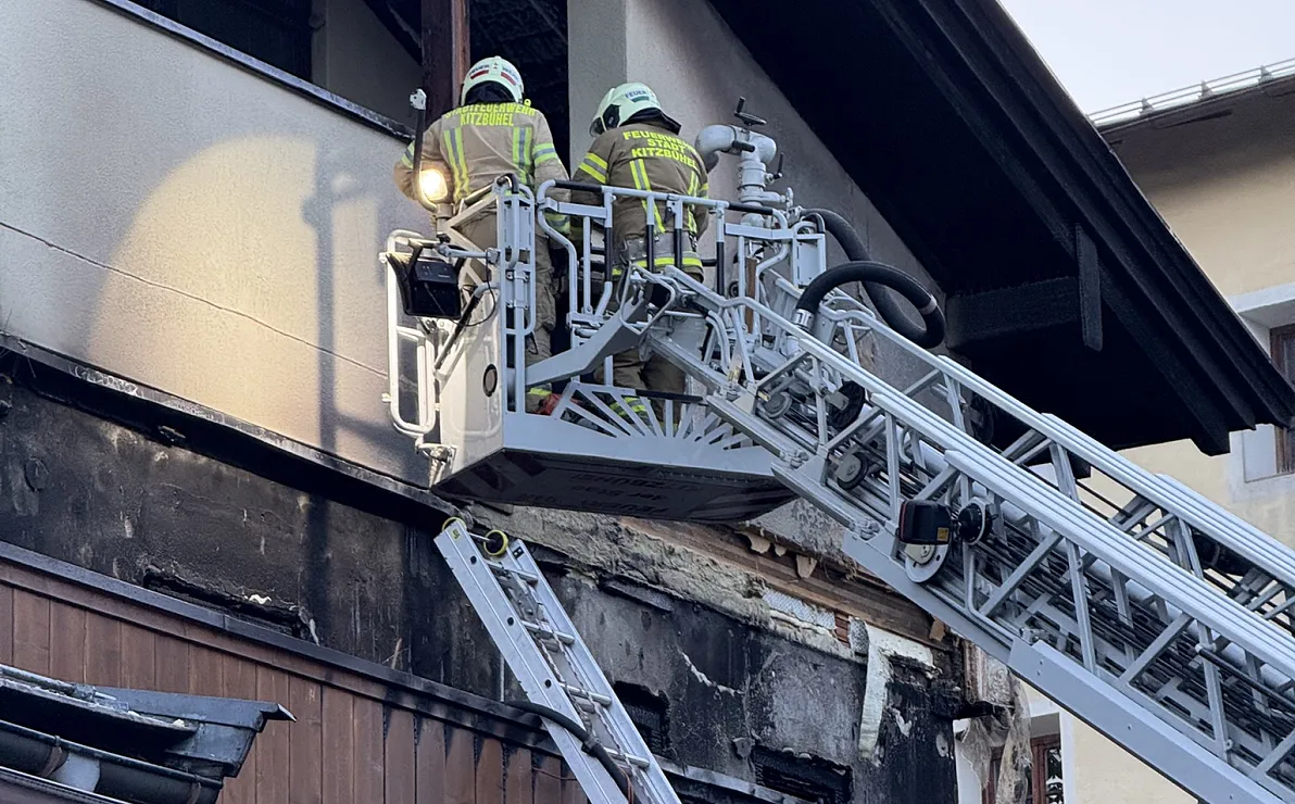 Zwei Feuerwehrleute in Schutzkleidung arbeiten von einem Hubsteiger aus an einem beschädigten Gebäude mit verrußter Fassade.