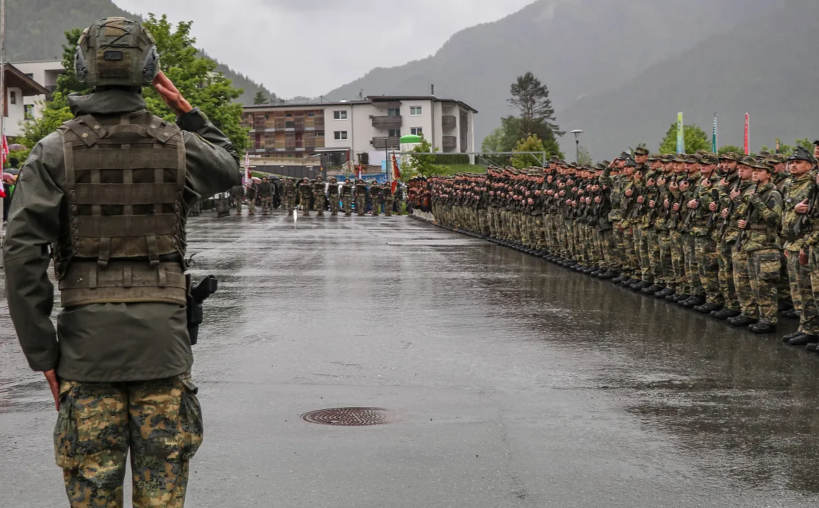 Soldaten in militärischer Formation, bei Regen auf einer nassen Straße stehend, während ein Offizier salutiert. Gebäude und Berge im Hintergrund.