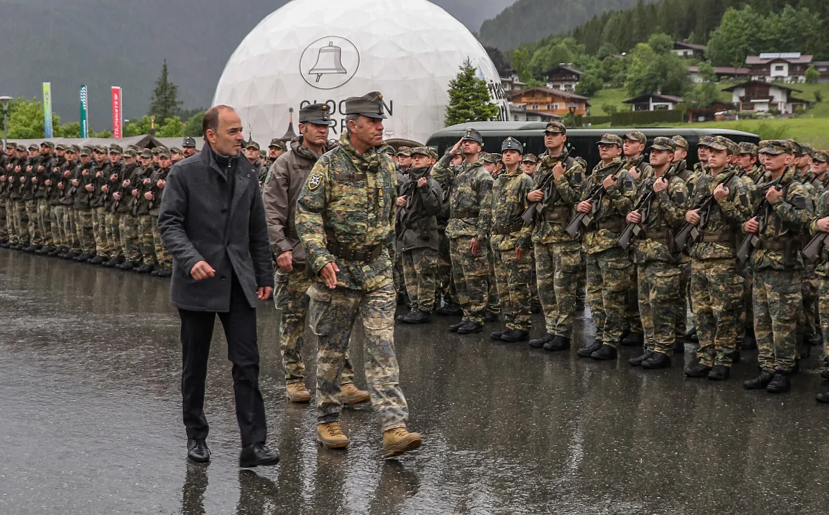 Zwei Männer schreiten vor einer Reihe von uniformierten Soldaten eine regennasse Straße entlang, im Hintergrund eine weiße Kuppel und Waldhütten.