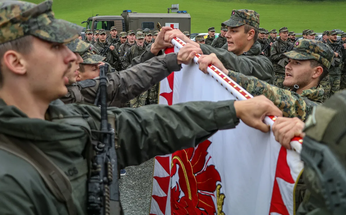 Soldaten in Militäruniform halten gemeinsam eine Fahne, während sie auf einem Feld stehen. Im Hintergrund sind weitere Soldaten und ein Militärfahrzeug zu sehen.