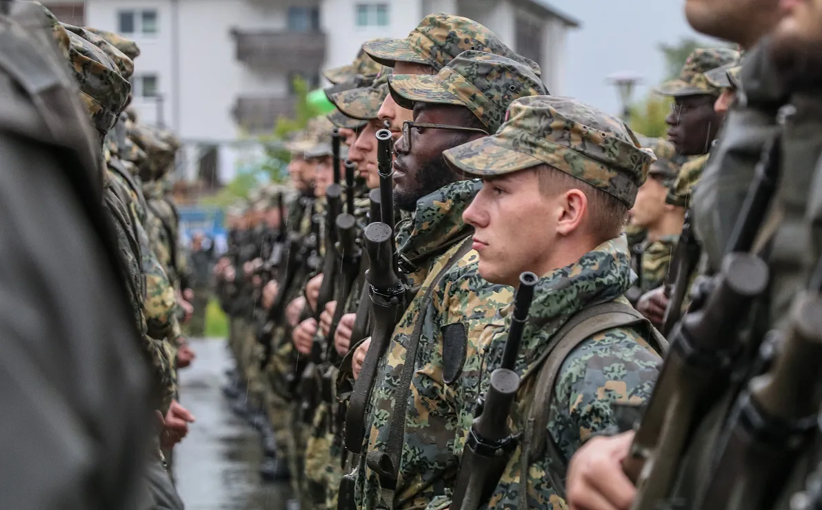 Soldaten in Tarnuniformen stehen in Reihen, mit Gewehren in der Hand. Im Hintergrund sind unscharfe Gebäude und Natur zu erkennen, die Szene wirkt ernst.