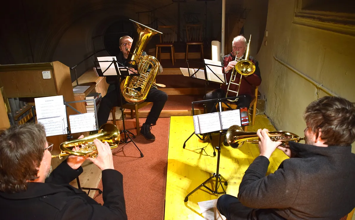 Vier Musiker spielen Blasinstrumente in einem kleinen Raum mit Notenständern. Sie tragen dunkle Kleidung und konzentrieren sich auf ihre Musiknoten.