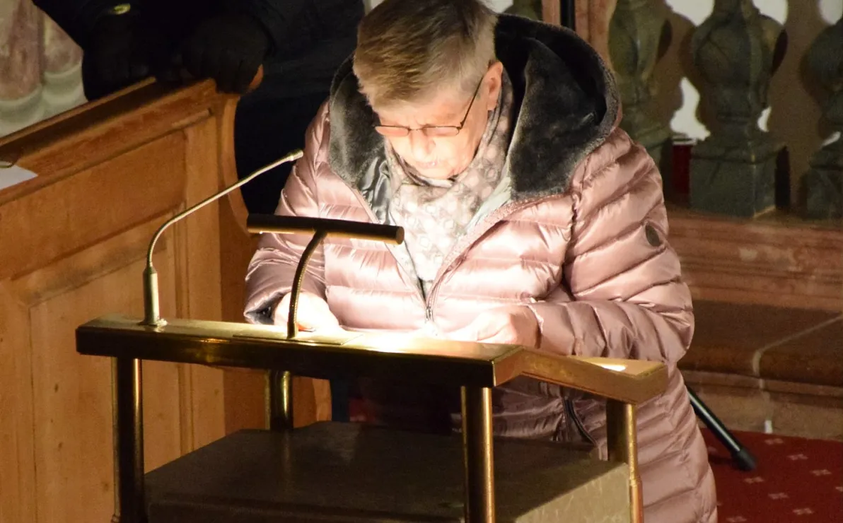 Ältere Person im Wintermantel steht an einem Rednerpult in einem Innenraum, liest scheinbar aus einem Buch vor. Der Raum wirkt warm beleuchtet.