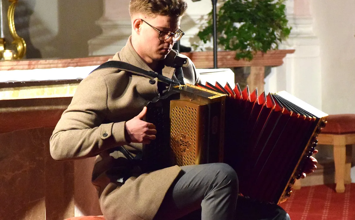 Mann spielt ein Akkordeon auf einem Sessel in einem dekorativen Raum mit Pflanzen im Hintergrund.