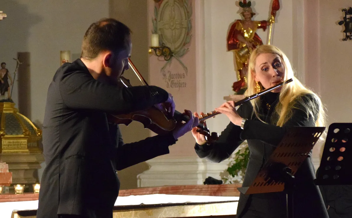 Ein Mann spielt Geige und eine Frau spielt Querflöte in einem Kircheninnenraum. Sie tragen schwarze Kleidung und stehen vor einem Altar mit Kerzen.