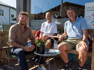 Drei Männer sitzen auf einer Terrasse vor einem Café, lächeln in die Kamera. Sie haben Getränke auf einem kleinen Tisch vor sich. Es ist sonniges Wetter.