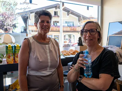 Zwei Frauen stehen in einem Raum mit Fenstern. Eine trägt ein ärmelloses Oberteil, die andere hält eine Wasserflasche. Im Hintergrund sind Snacks und Pflanzen.