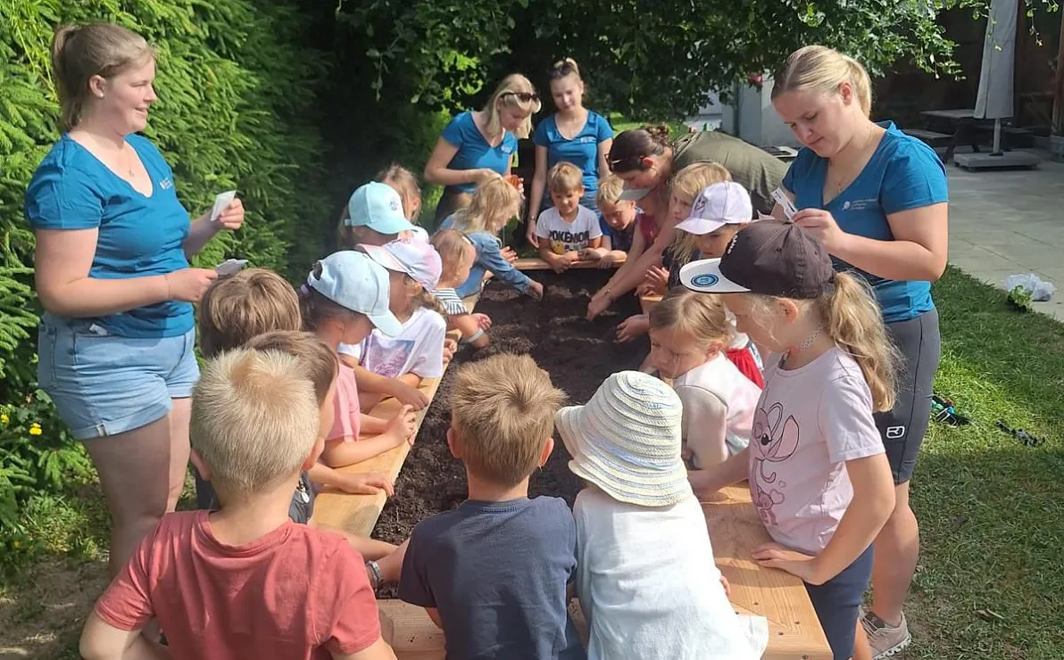 Kinder und Erwachsene pflanzen gemeinsam in einem Garten. Sie arbeiten um ein großes Hochbeet herum, umgeben von Bäumen und grüner Vegetation.