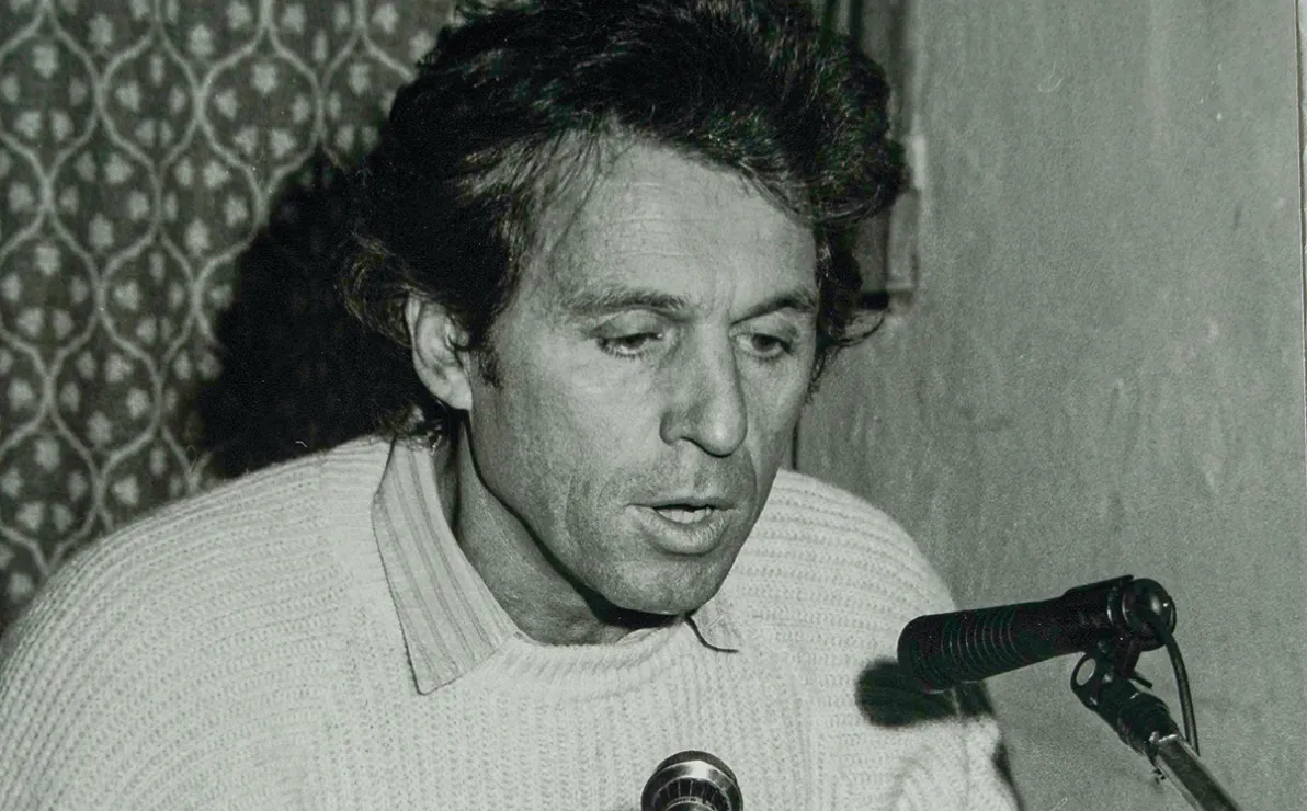 Ein Mann mit lockigem Haar spricht in ein Mikrofon. Er trägt einen Strickpullover und sitzt hinter einem Tisch mit weiterem Mikrofonequipment.