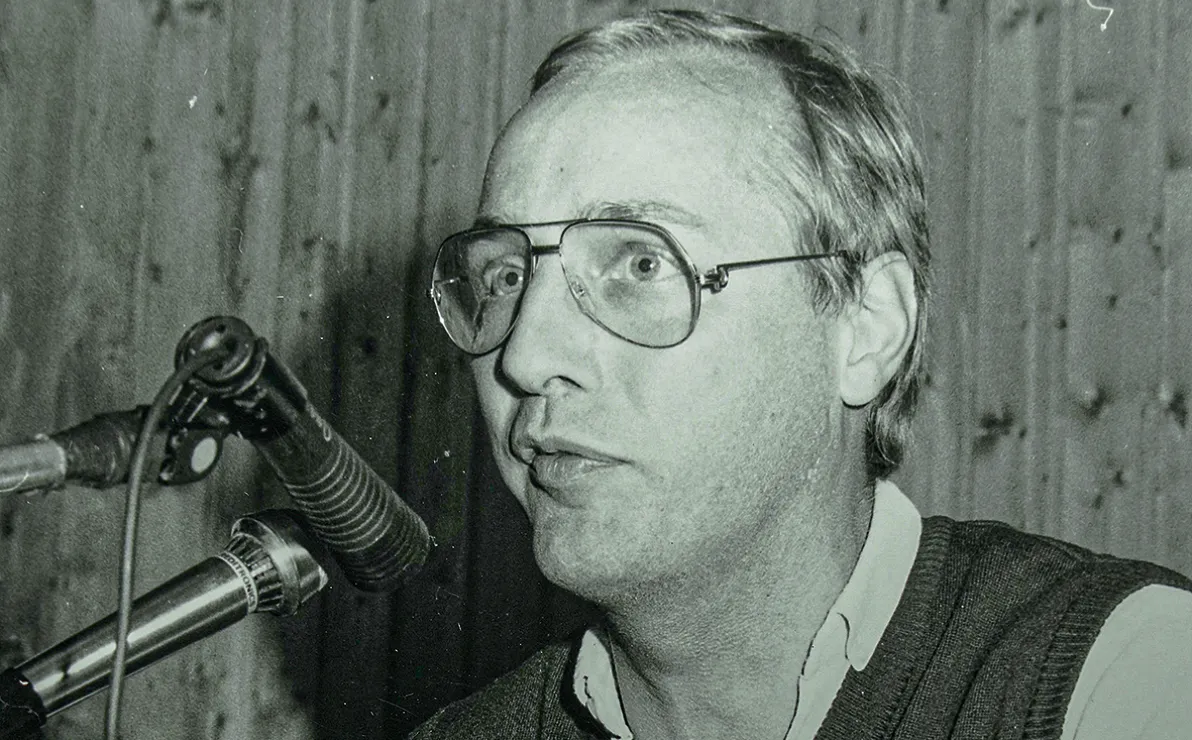Ein Mann mit Brille und Weste spricht in ein Mikrofon. Der Hintergrund ist holzvertäfelt, das Bild ist in Schwarz-Weiß gehalten.