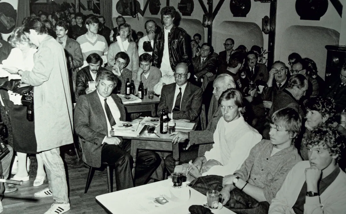 Ein schwarz-weiß Foto zeigt eine lebhafte Bar oder Clubszene. Menschen sitzen an überfüllten Tischen, während einige stehen oder sich unterhalten. Stimmung: gesellig.