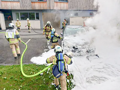 Feuerwehrleute bekämpfen einen Fahrzeugbrand mit Schaum, während dichter Rauch aufsteigt. Ein Team arbeitet konzentriert vor einem Gebäude.
