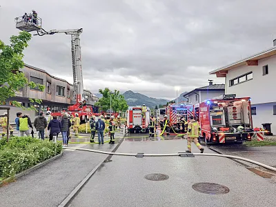 Feuerwehreinsatz auf einer Straße mit mehreren Löschfahrzeugen und Feuerwehrleuten. Ein Leiterwagen ist ausgefahren, Menschen beobachten die Szene.