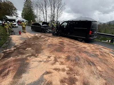 Ein Autounfall auf einer Landstraße mit zwei Fahrzeugen und Feuerwehrkräften, die die Unfallstelle sichern. Der Asphalt ist mit einem Material bestreut.