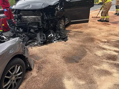 Ein Autounfall mit zwei beschädigten Fahrzeugen auf einer Straße. Feuerwehrkräfte sind vor Ort, um Hilfe zu leisten. Der Boden ist mit Sand bedeckt.