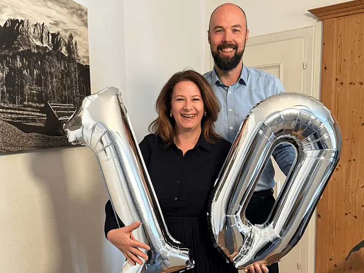 Zwei lächelnde Personen halten silberne Luftballons in Form der Zahl 10 in einem Raum mit einem Bild im Hintergrund.