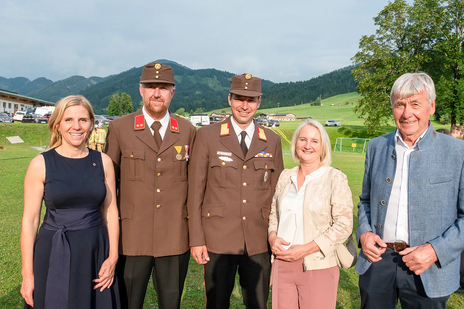 Fünf Personen stehen auf einer Wiese vor einer Bergkulisse, zwei tragen Uniformen, die anderen sind in Freizeitkleidung. Es scheint eine Veranstaltung zu sein.