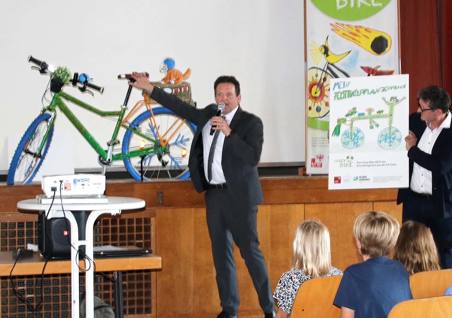 Zwei Männer präsentieren auf einer Bühne ein grünes Fahrrad und ein Plakat vor einem Publikum. Kinder sitzen zu und sehen interessiert zu.