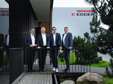 Vier Männer in Anzügen stehen auf einer Terrasse vor einem modernen Gebäude mit dem Schriftzug 'Egger'. Im Hintergrund sind Bäume und Pflanzen sichtbar.