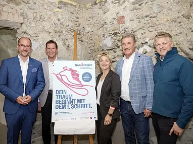 Gruppe von fünf Personen steht vor einem Plakat mit der Aufschrift: "Dein Traum beginnt mit dem 1. Schritt". Hintergrund besteht aus Steinwänden.