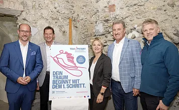 Gruppe von fünf Personen steht vor einem Plakat mit der Aufschrift: "Dein Traum beginnt mit dem 1. Schritt". Hintergrund besteht aus Steinwänden.