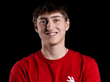 Ein junger Mann trägt ein rotes T-Shirt und lächelt vor einem schwarzen Hintergrund. Er hat kurzes, braunes Haar und verschränkte Arme.