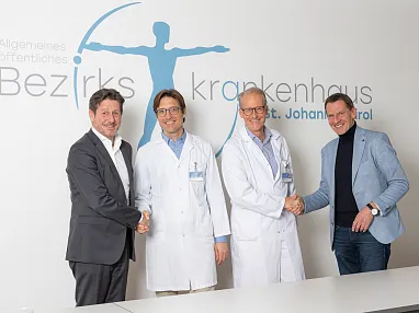 Vier Männer, zwei in weißen Kitteln, schütteln sich vor einem Krankenhausplakat die Hände. Sie lächeln und stehen nah beieinander.