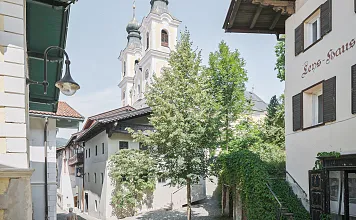 Historische Altstadtgasse mit malerischen Gebäuden und einer Kirche im Hintergrund. Grüne Bäume säumen die Straße an einem sonnigen Tag.