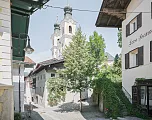 Historische Altstadtgasse mit malerischen Gebäuden und einer Kirche im Hintergrund. Grüne Bäume säumen die Straße an einem sonnigen Tag.