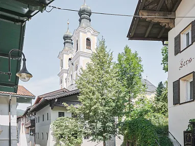 Historische Altstadtgasse mit malerischen Gebäuden und einer Kirche im Hintergrund. Grüne Bäume säumen die Straße an einem sonnigen Tag.