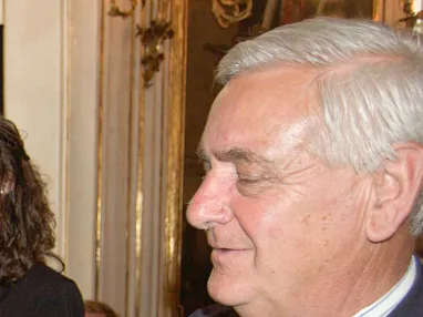 Drei Personen bei einer Zeremonie in einem festlichen Raum, zwei halten Auszeichnungen. Ein Mann im Anzug lächelt, während ihm ein Preis überreicht wird.