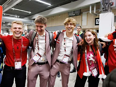 Eine Gruppe junger Menschen in festlicher Kleidung steht lächelnd mit den Armen um die Schultern und freut sich in einem belebten Veranstaltungsraum.