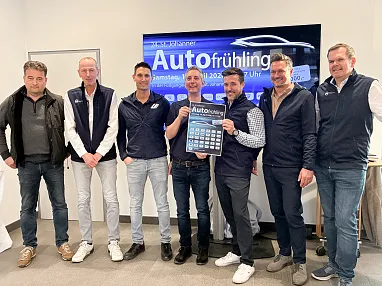 Sechs Männer stehen vor einem Werbebanner für eine Automesse. Zwei halten ein Plakat, während die anderen lächeln. Alle tragen legere Kleidung.