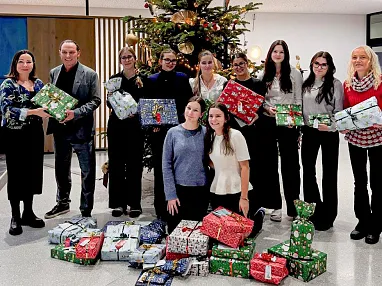 Gruppe von Menschen steht lächelnd vor einem großen, geschmückten Weihnachtsbaum und hält bunte Geschenke in den Händen.
