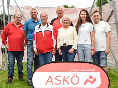 Eine Gruppe von sieben Personen steht lächelnd zusammen vor einem ASKÖ-Schild auf einer Wiese. Sie tragen Freizeitkleidung in rot-weißen Farbtönen.