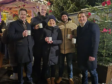 Fünf Personen in Winterkleidung stehen mit Tassen vor einem geschmückten Weihnachtsbaum auf einem Weihnachtsmarkt, umgeben von Lichtern und Dekorationen.