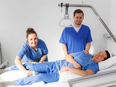 Zwei Personen in blauen Krankenhauskleidern stehen neben einem Patienten in einem Bett in einem Krankenhaussaal und lächeln in die Kamera.