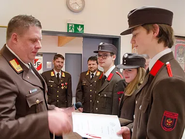 Eine Gruppe von Personen in Uniform mit Abzeichen steht in einem Raum. Eine Person überreicht einer anderen ein Dokument. Ein Gruppenfoto ist im Hintergrund erkennbar.