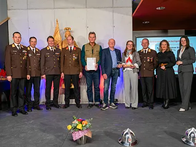 Eine Gruppe von Menschen steht auf einer Bühne, einige in Uniform, andere in Zivilkleidung. Sie halten Zertifikate und Trophäen, Blumen sind am Boden drapiert.