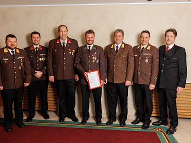 Zwei lächelnde Männer in dunklen Uniformen mit Abzeichen und Medaillen vor einem neutralen Hintergrund, möglicherweise Mitglieder einer Organisation.