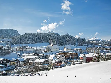 Verschneite Berglandschaft mit einem malerischen Dorf, Kirche im Zentrum, umliegende Wälder und Berge unter blauem Himmel mit ein paar Wolken.