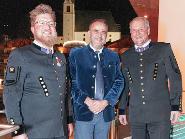 Drei Männer stehen lächelnd nebeneinander. Zwei tragen dunkle Uniformen mit Abzeichen, der Mann in der Mitte trägt eine blaue Jacke und eine Krawatte.
