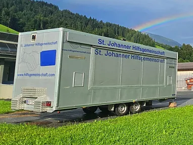 Ein Regenbogen spannt sich über einer grünen Wiese und einem Gebäude, während ein grauer LKW-Kastenanhänger auf einer asphaltierten Fläche steht.
