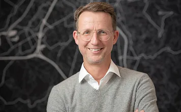 Ein Mann mit Brille und grauem Pullover steht lächelnd vor einer Wand mit abstraktem Linienmuster. Er hat braune Haare und trägt ein weißes Hemd darunter.
