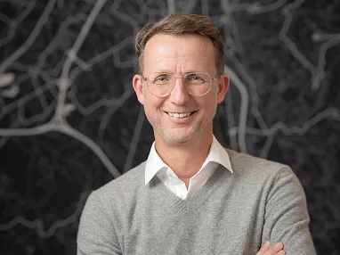 Ein Mann mit Brille und grauem Pullover steht lächelnd vor einer Wand mit abstraktem Linienmuster. Er hat braune Haare und trägt ein weißes Hemd darunter.