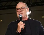 Mann mit Brille und Mikrofon spricht auf einer Bühne, trägt ein dunkles Jackett, dunkles Hemd und steht vor einem beleuchteten Hintergrund.